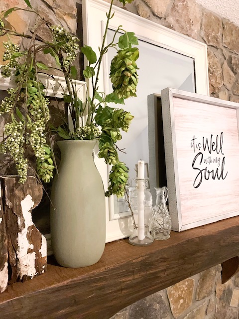 A cottage style mantel decorated for spring with faux greenery, a mirror, a picture, and empty frames