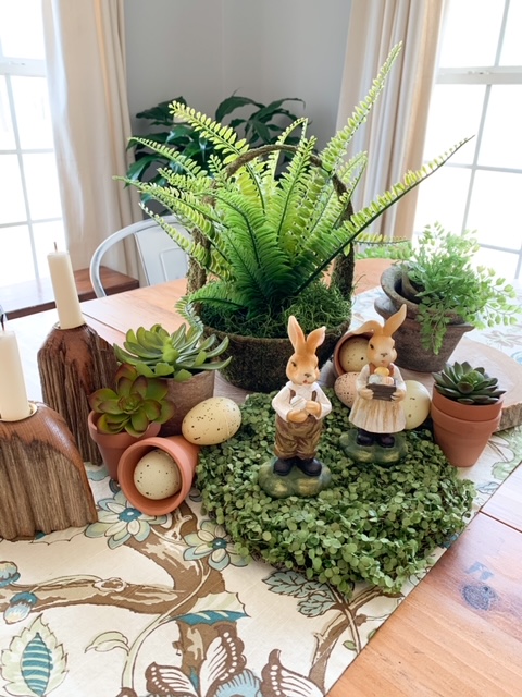 A spring table centerpiece made from faux greenery, wood rounds, wood candlesticks, faux eggs, and bunnies