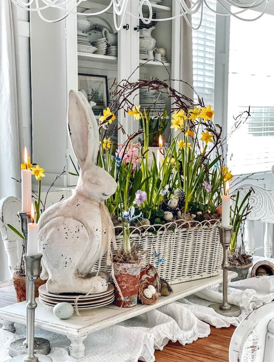 a white wicker baskets holding spring flowers surrounded by a white concrete bunny, candles, and bird nest