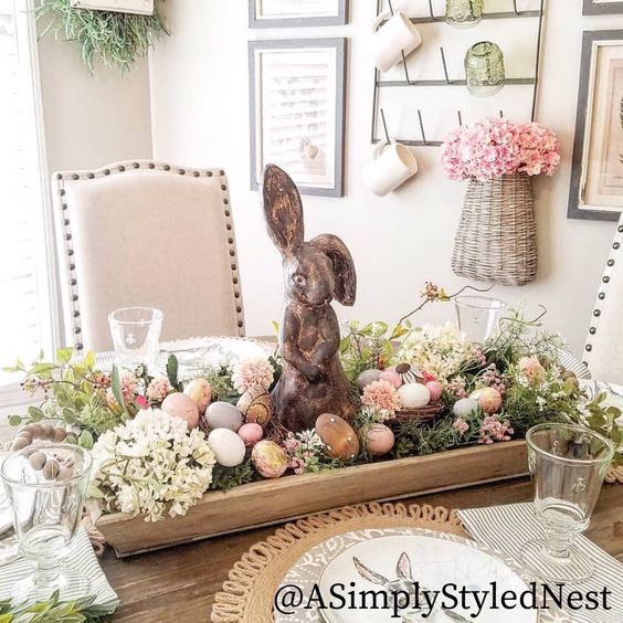 wood tray in the center of a table holding a concrete bunny figurine, faux eggs, and spring forals